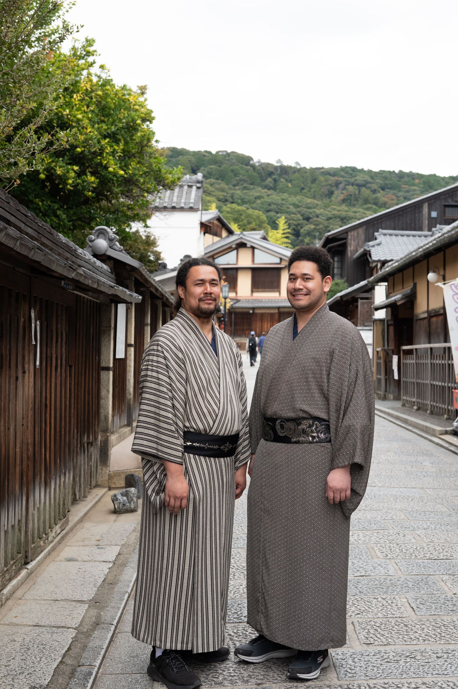 Two men in traditional Japanese yukata pose together on historic Kyoto street with forested hills.