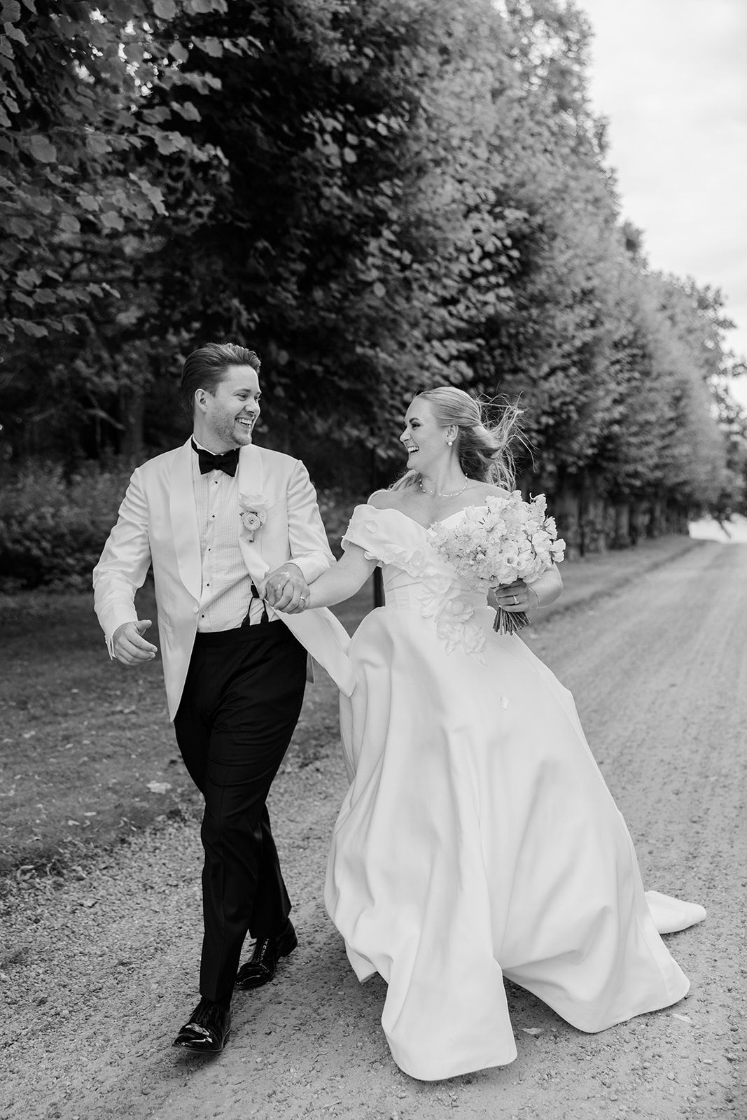 A bride and groom share a joyful moment while walking down a tree-lined path in black and white photography.