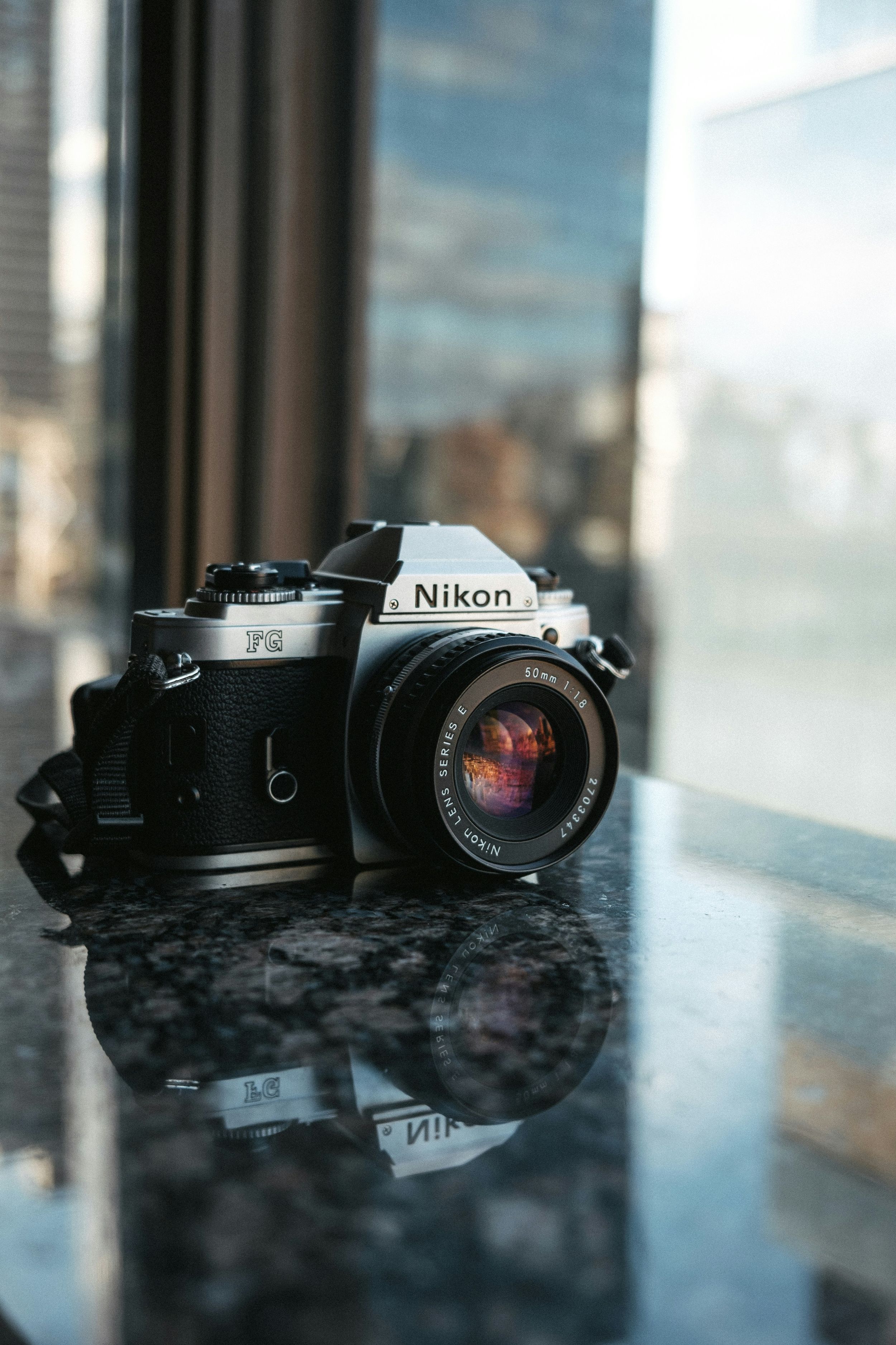 A black Nikon film camera with lens displayed on a reflective surface near a window.