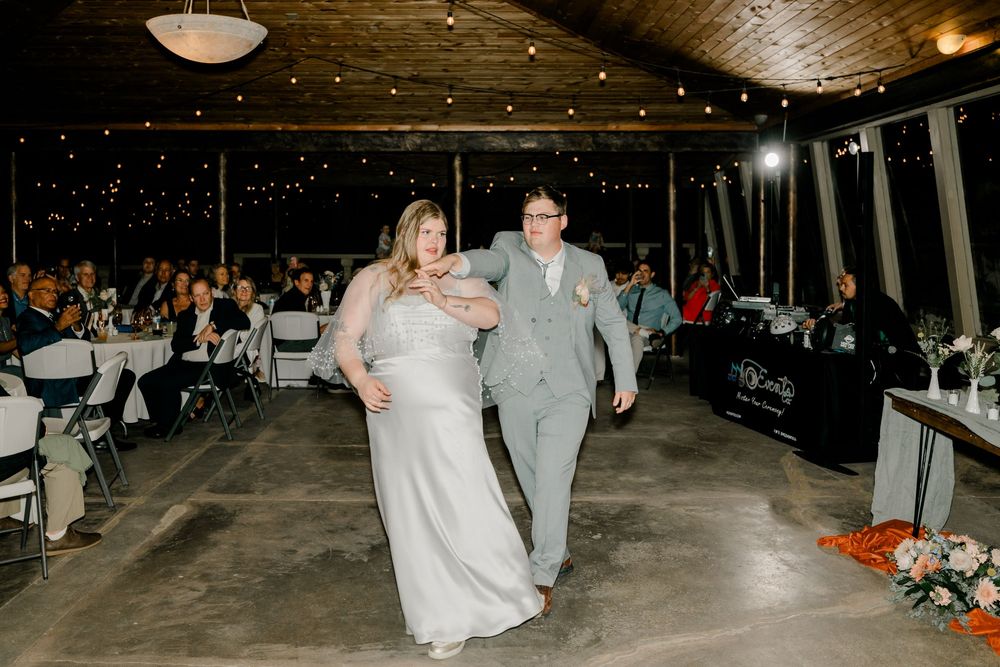 Wedding guests dance and celebrate on a rustic wooden dance floor under warm string lights at an evening reception.
