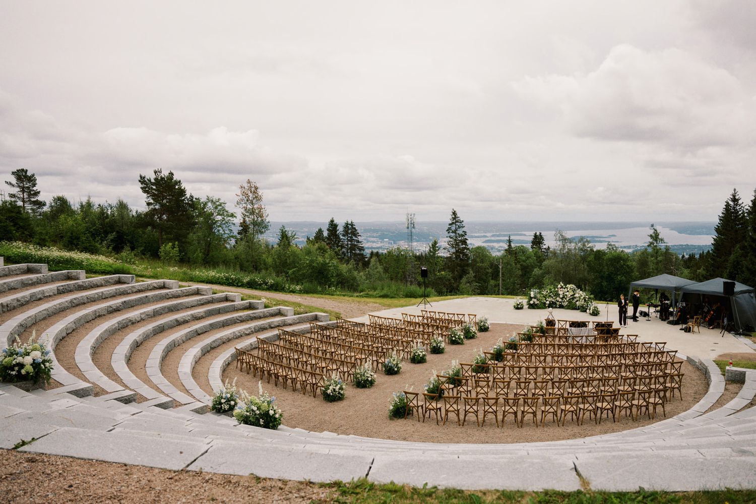 An outdoor amphitheater with curved stone seating and wooden chairs arranged for a ceremony overlooking a forested landscape.