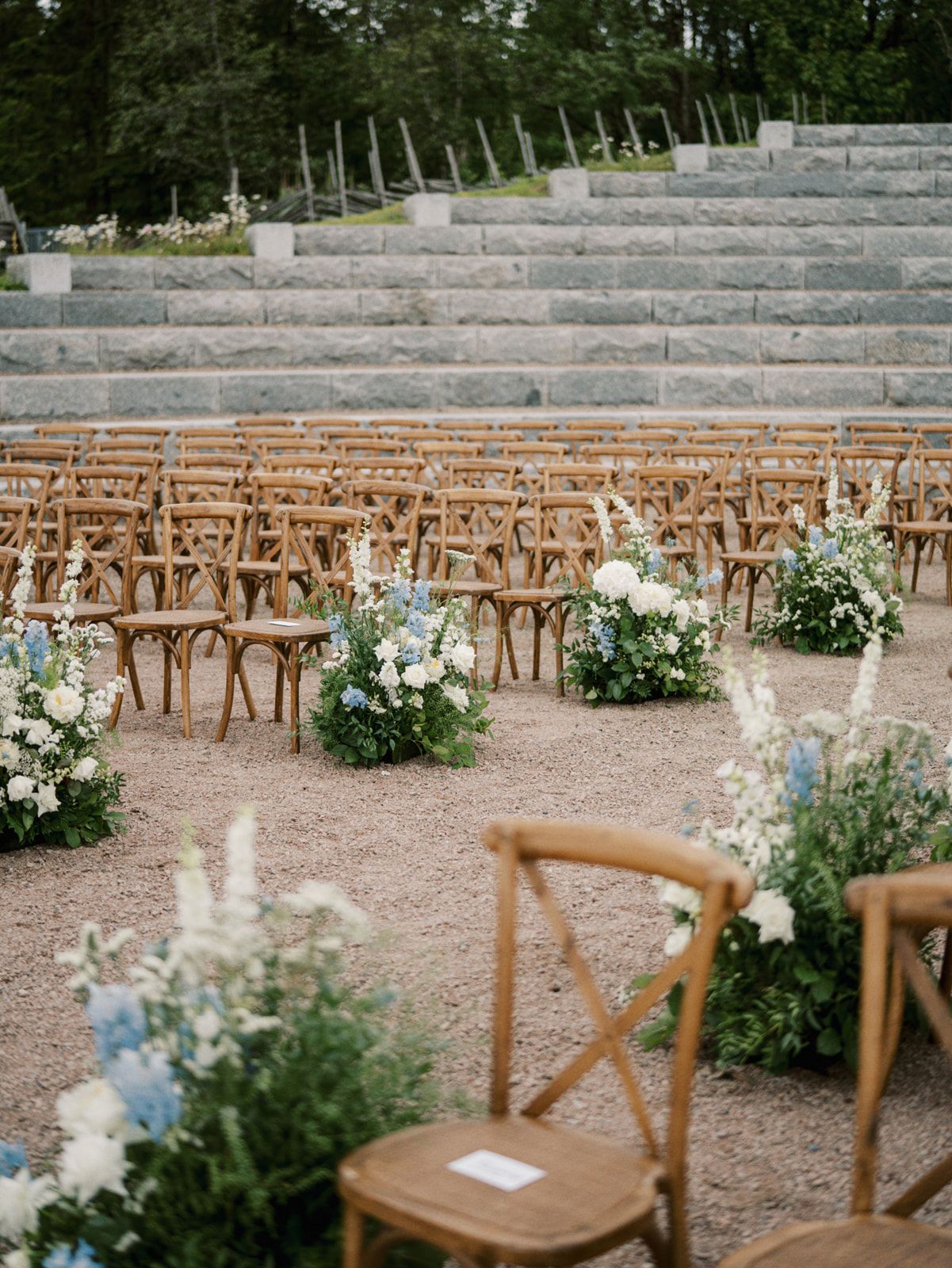 Rustic outdoor wedding ceremony setup with wooden cross-back chairs and stone amphitheater steps decorated with white floral arrangements.