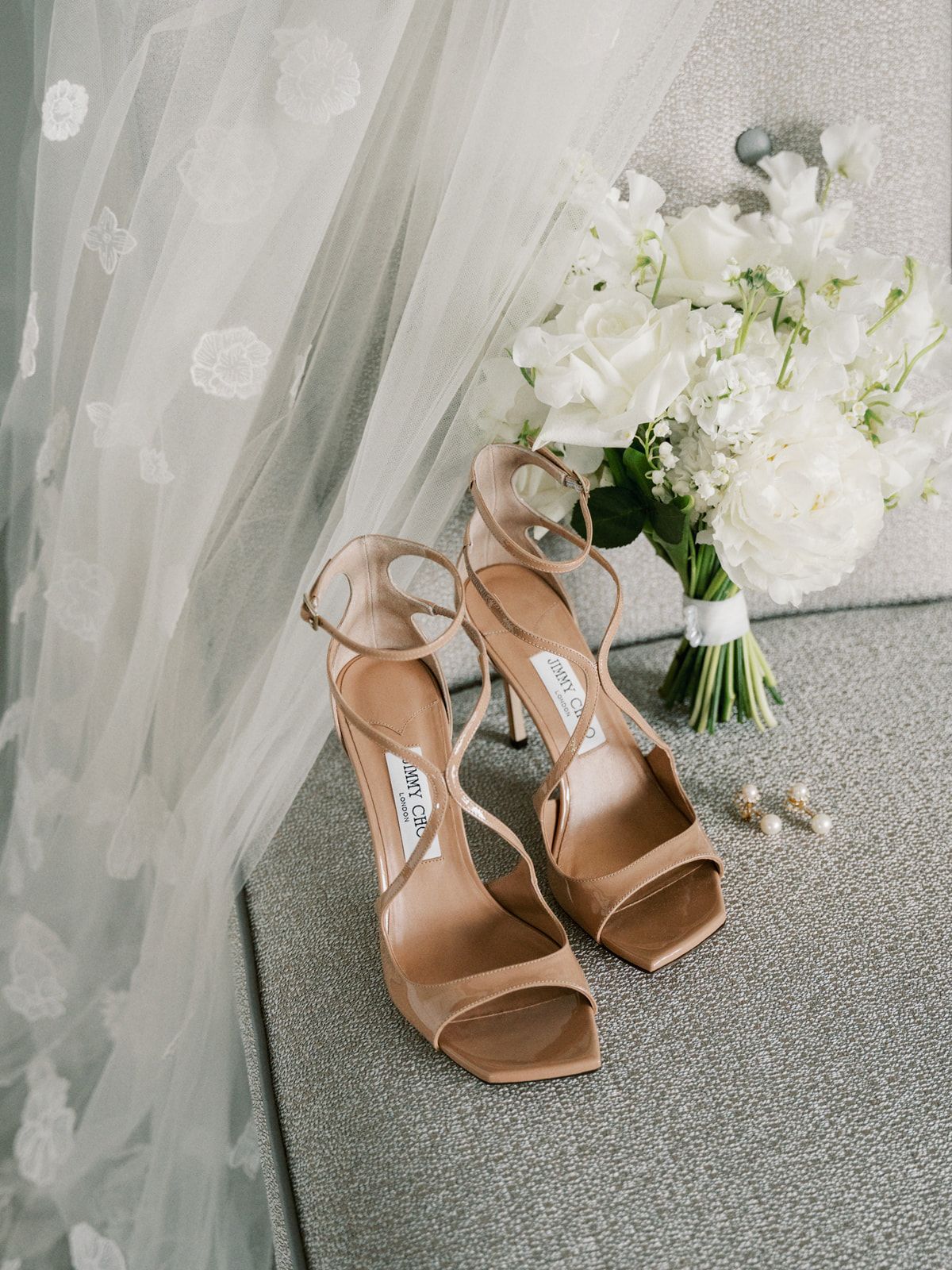 Elegant nude strappy heels placed beside a white floral bouquet against a sheer curtain background.