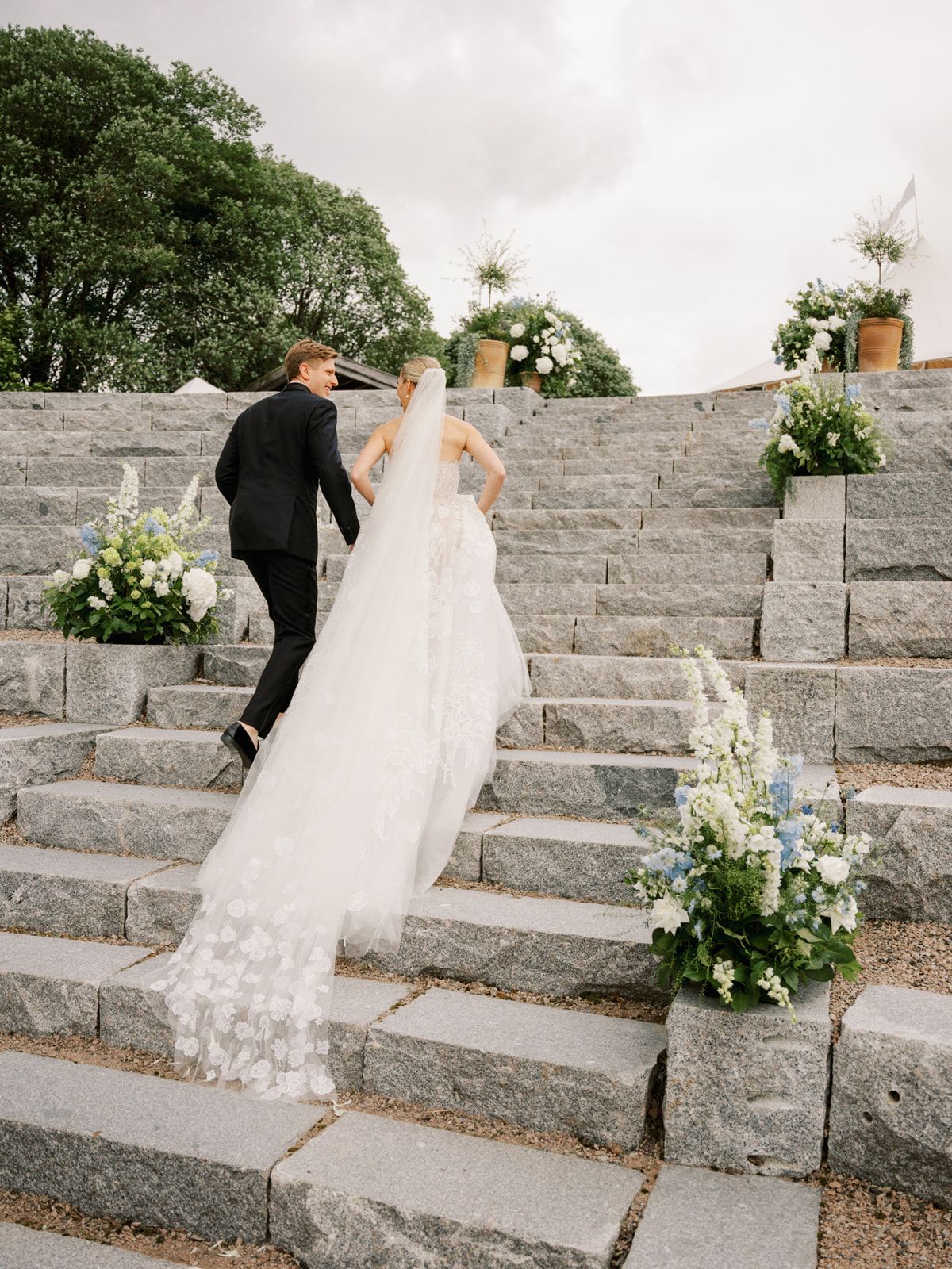 A couple climbs stone steps decorated with white floral arrangements and potted plants during an outdoor wedding ceremony.