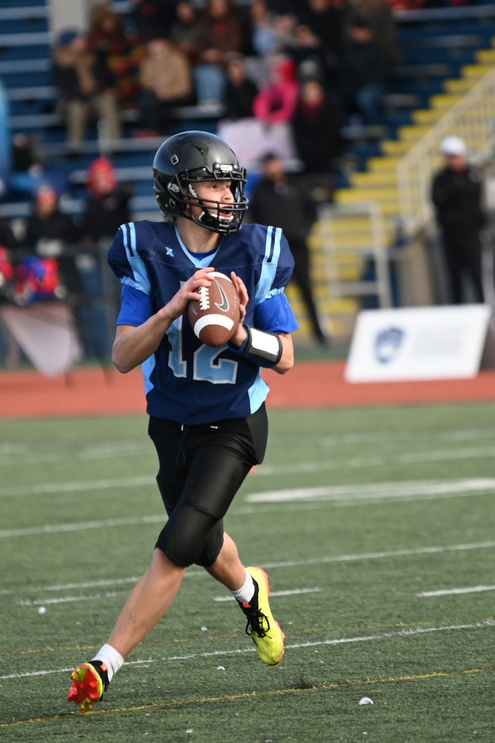 Football player in blue jersey and black pants prepares to throw the ball during a game on artificial turf.