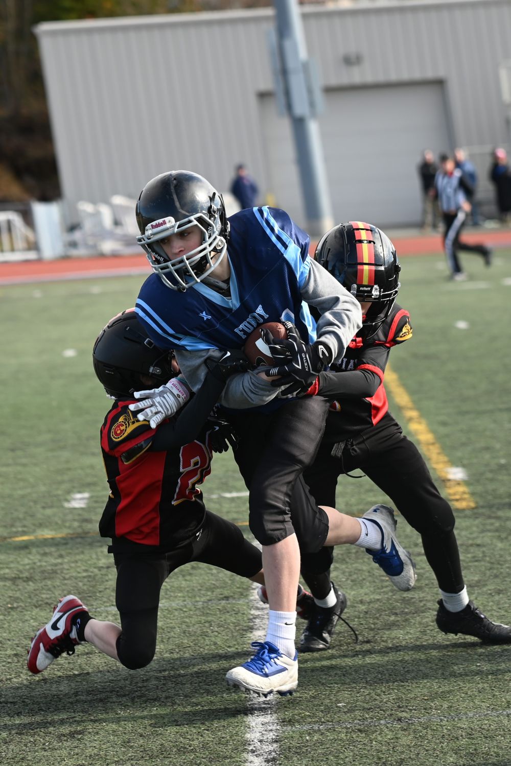 Youth football players in black and red uniforms compete during a tackle play on an artificial turf field.