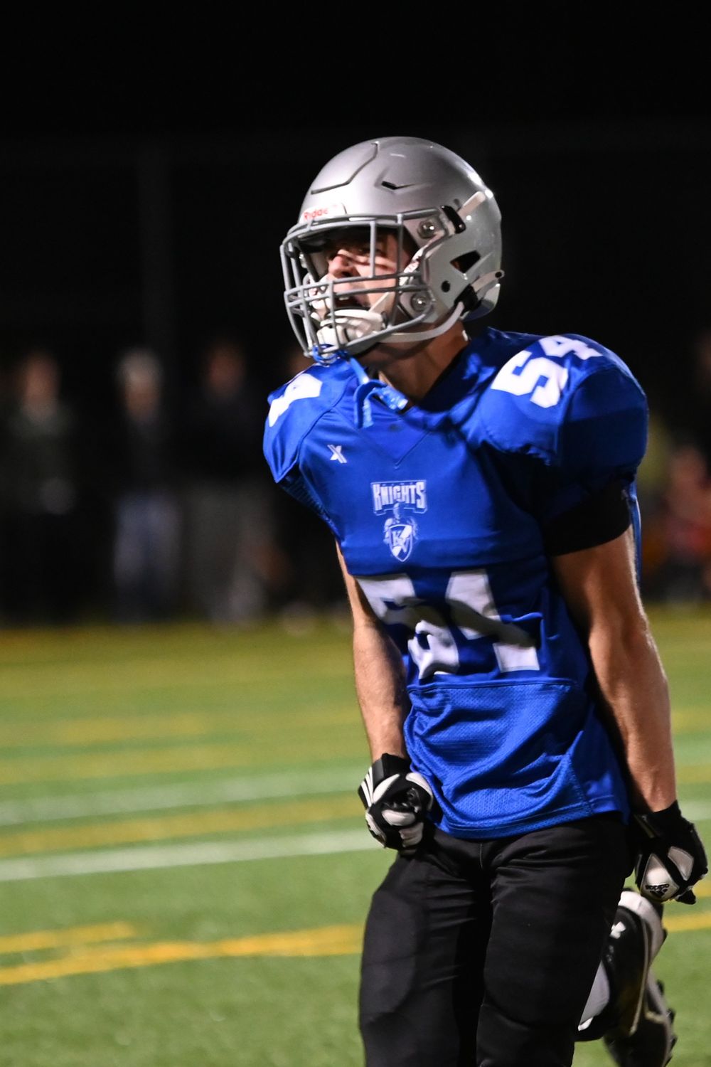Football player in blue jersey and black pants crouches in defensive stance during nighttime game.