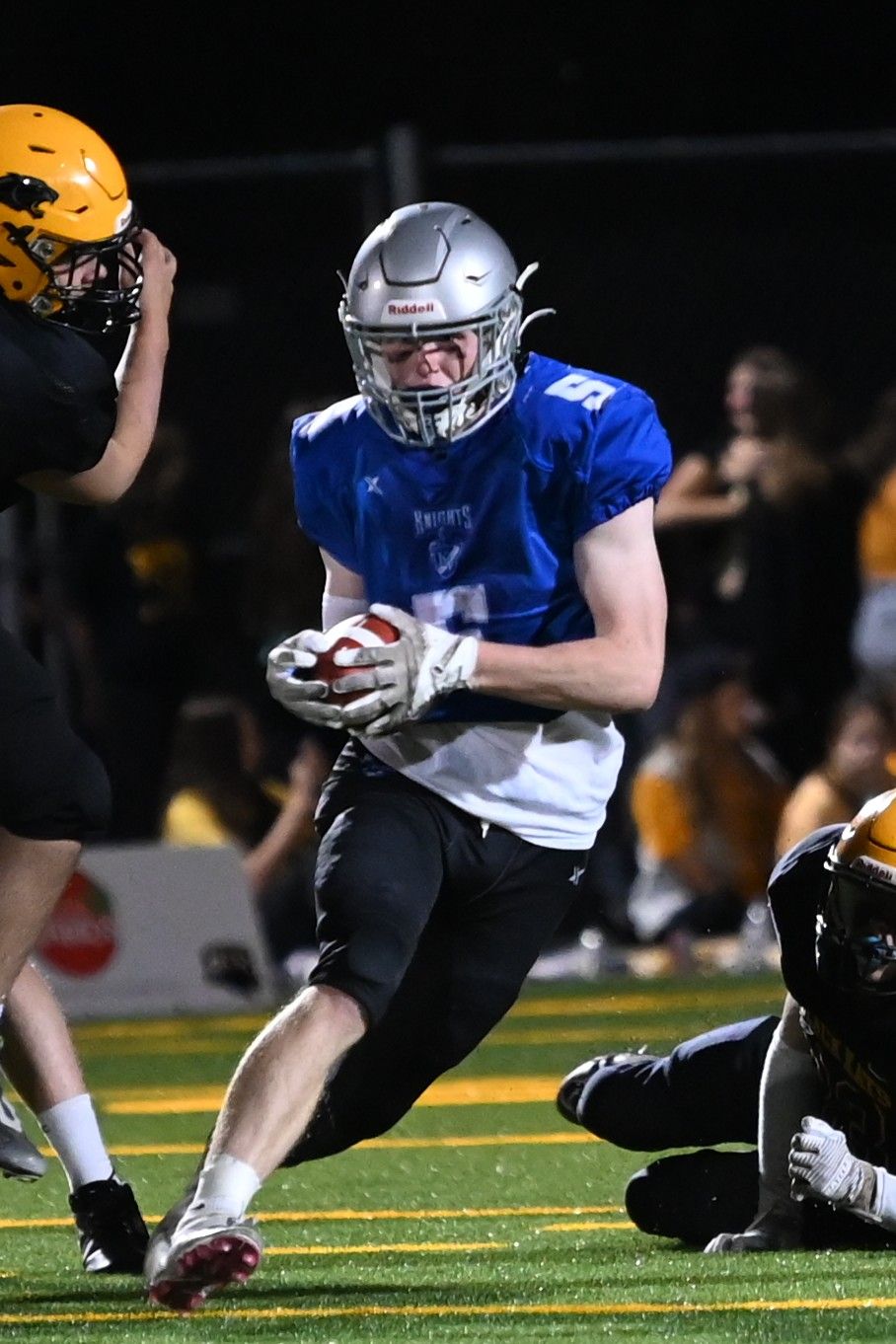 Football players in blue and white uniforms dive and tackle during an intense nighttime game on artificial turf.