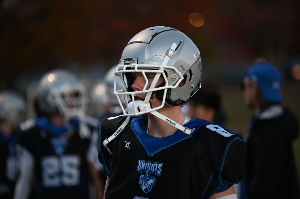 Football players wearing silver helmets and uniforms stand on the sidelines during a game.