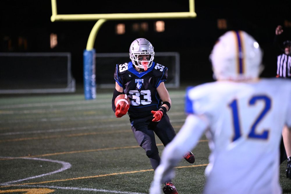 Football player in dark jersey runs with ball during nighttime game on illuminated field.