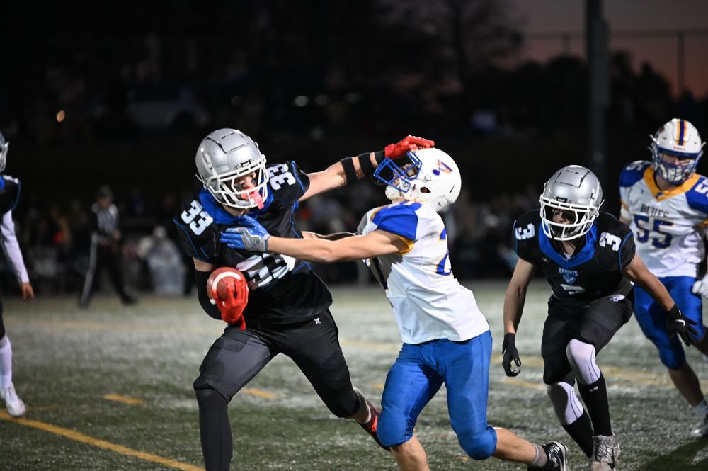 Football players from opposing teams collide during a tackle in a high school game on the field at night.