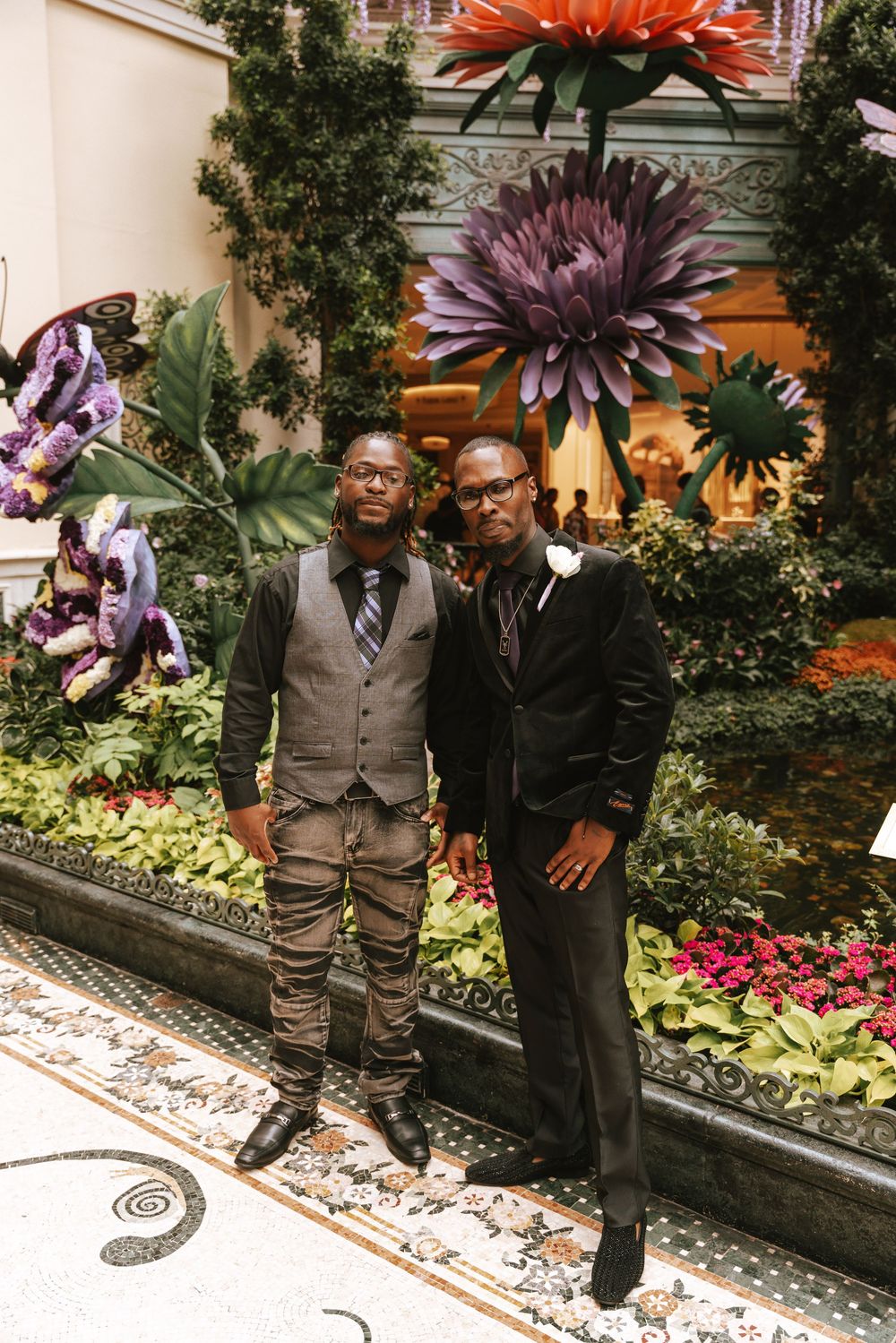 Two well-dressed men pose in front of a garden display with large decorative flowers at the Bellagio Conservatory.