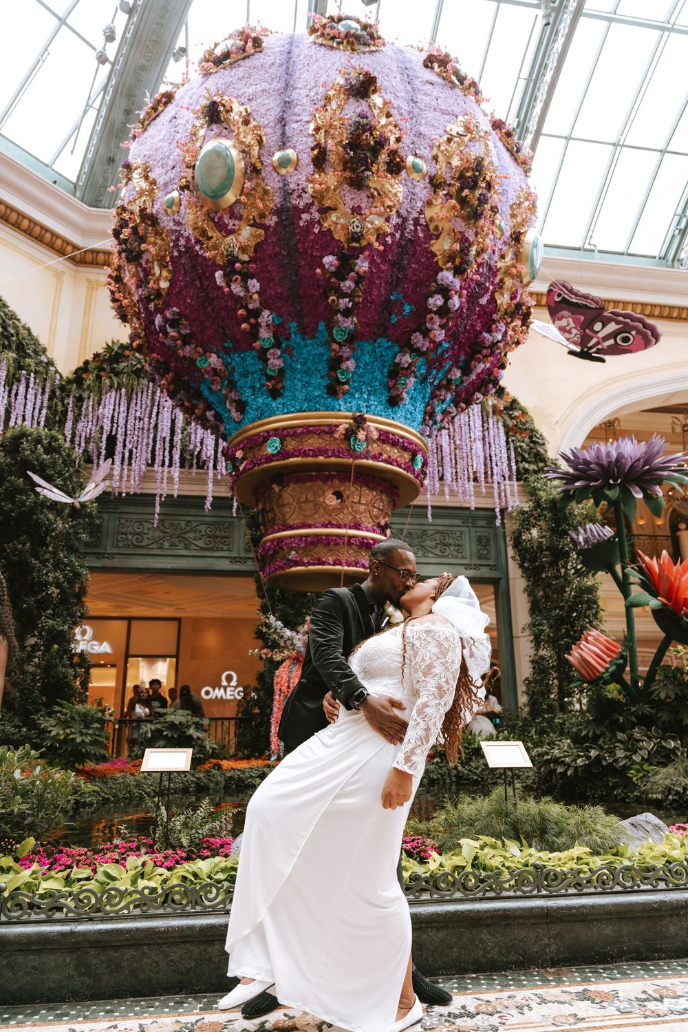 A person in a white dress spins and twirls through an ornate indoor garden conservatory with hanging floral decorations.