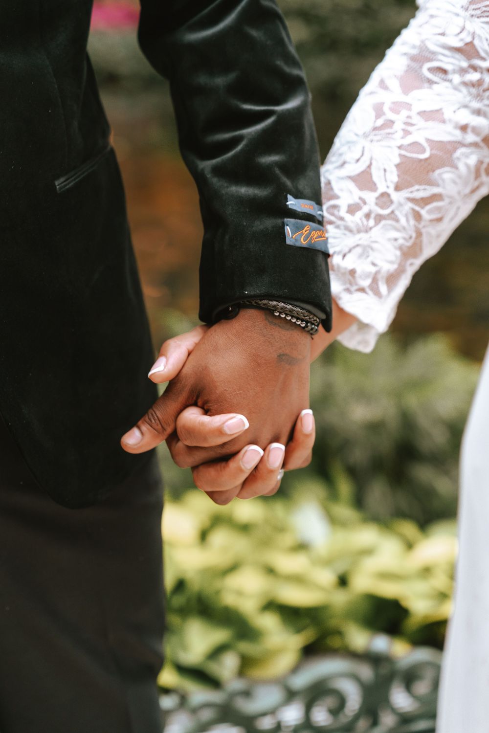 Close-up photo of a couple holding hands with delicate lace sleeve detail against a blurred natural background.