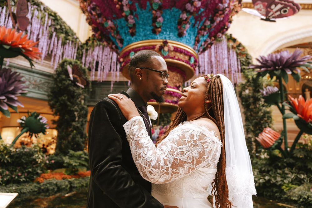 A couple shares an intimate moment at their wedding ceremony under colorful floral decorations and hanging crystals.