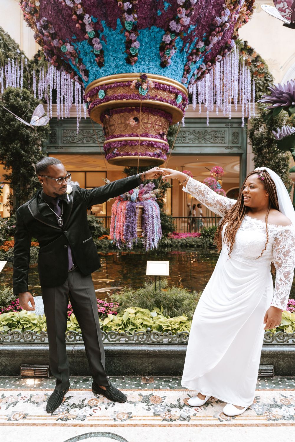 A couple performs a joyful dance sequence in front of a garden backdrop, with the bride in white and groom in black.