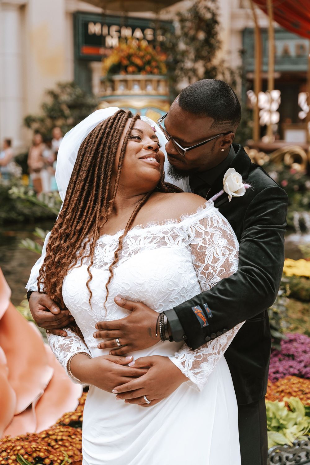 Newlywed couple embraces in front of floral display at outdoor wedding venue, bride wearing lace gown and groom in black suit.