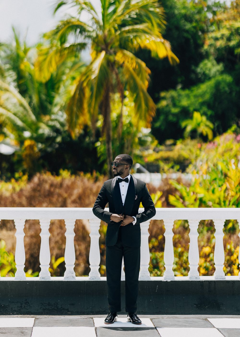 A person in a black tuxedo and bow tie stands on a white balcony with lush tropical foliage in the background.