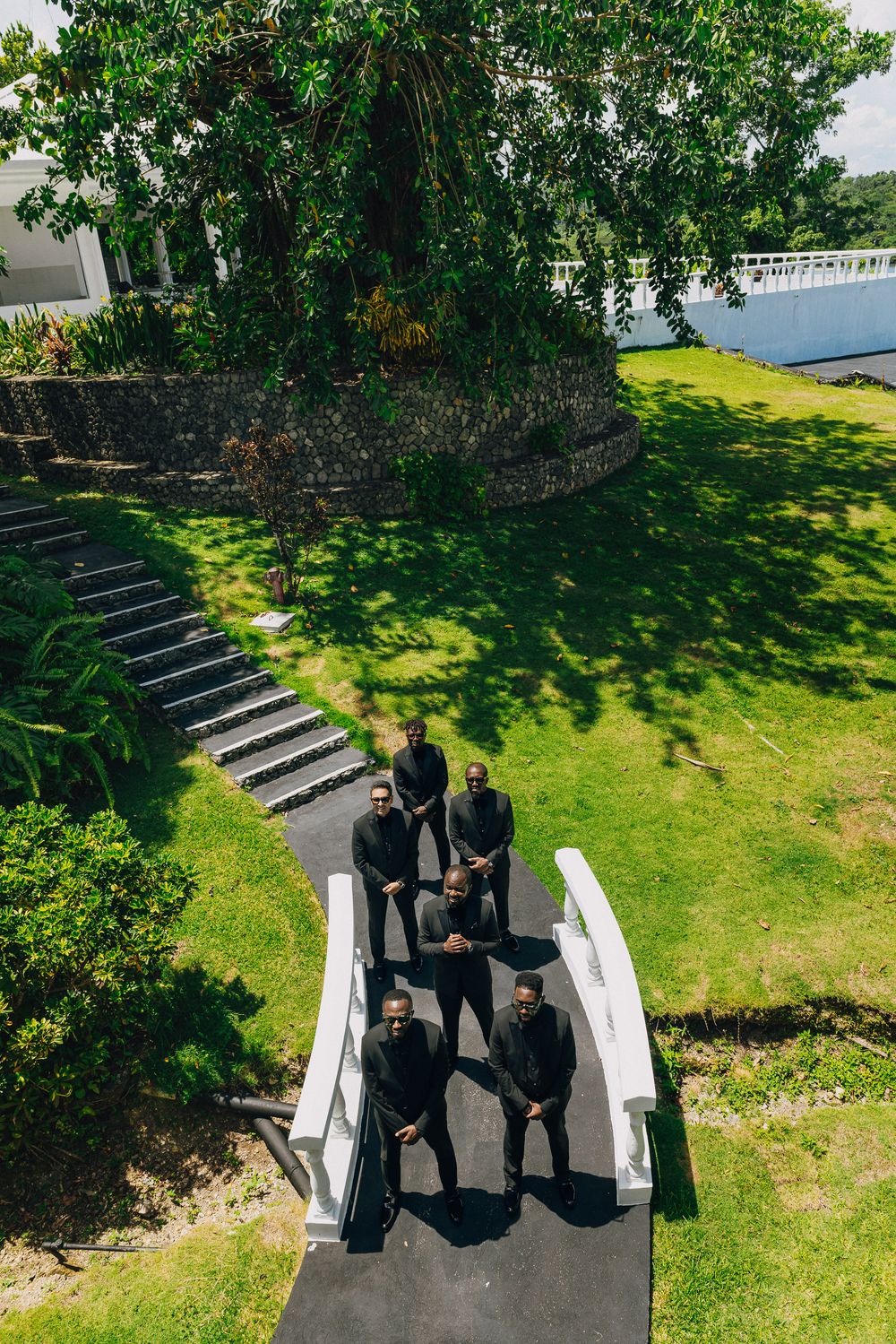 Aerial view of people in formal attire walking along a white pathway surrounded by lush green lawn and trees.