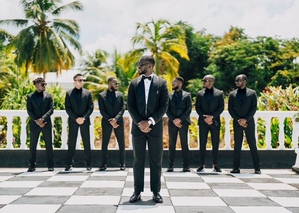 Wedding party members in black suits and dresses stand in a line against a tropical backdrop of palm trees.