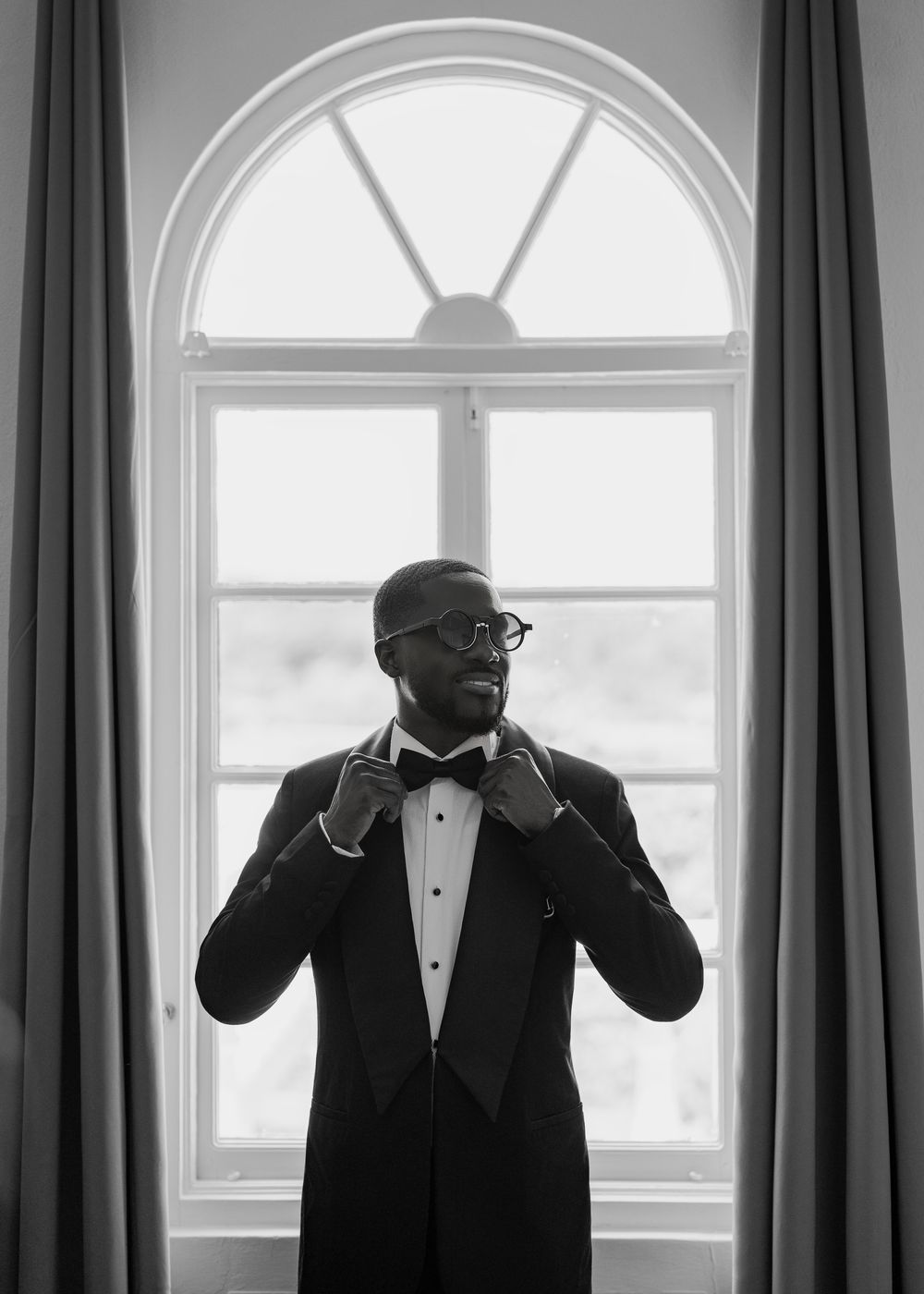 Three black and white photos show a person in a tuxedo adjusting their bowtie near elegant arched windows.
