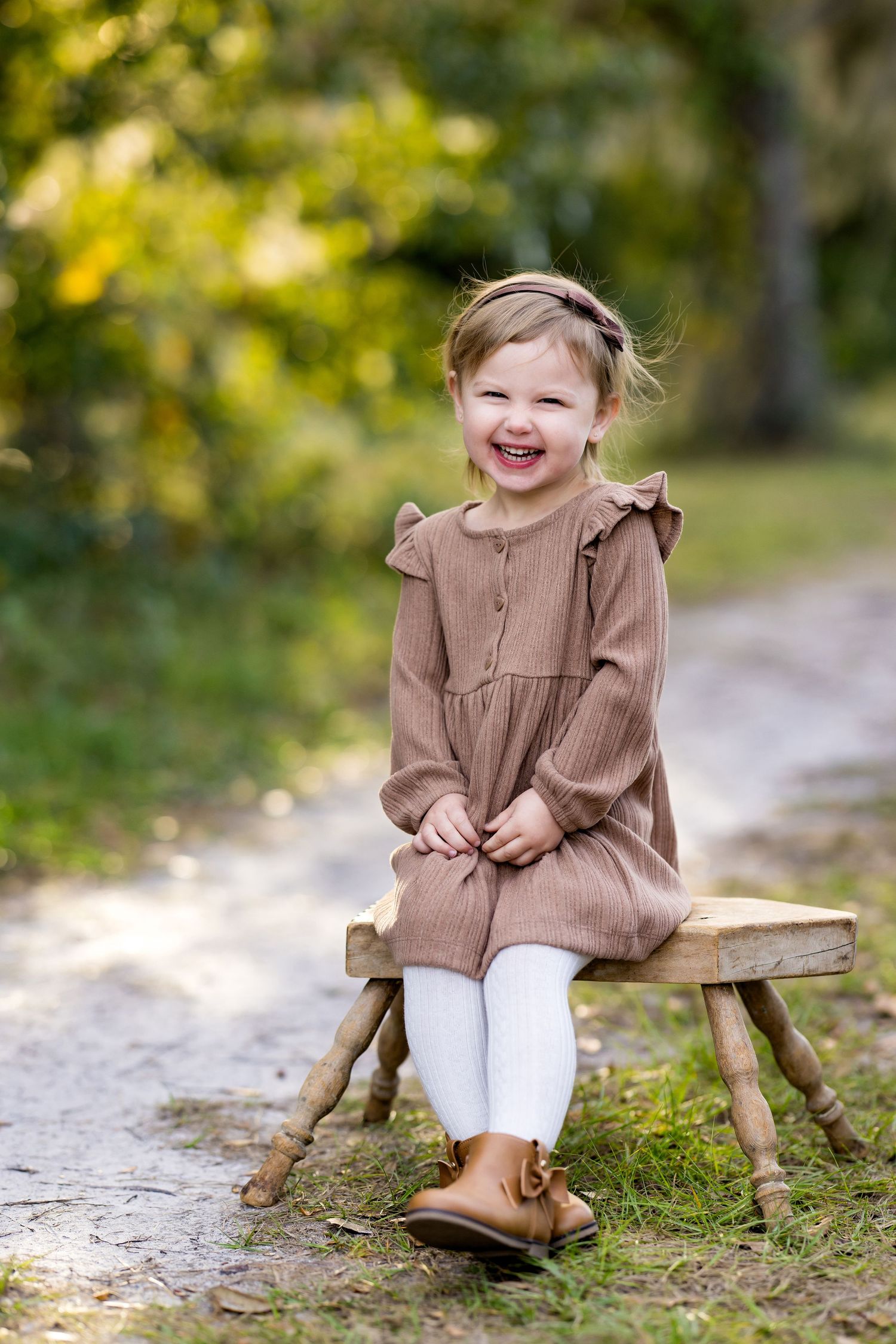 Child in brown ruffle dress and white leggings sits on wooden log in outdoor autumn setting.