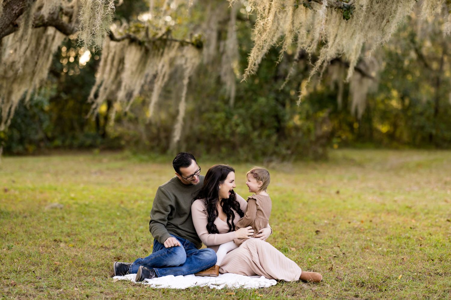 Family sits together on blanket in grass field with Spanish moss hanging from trees in background.