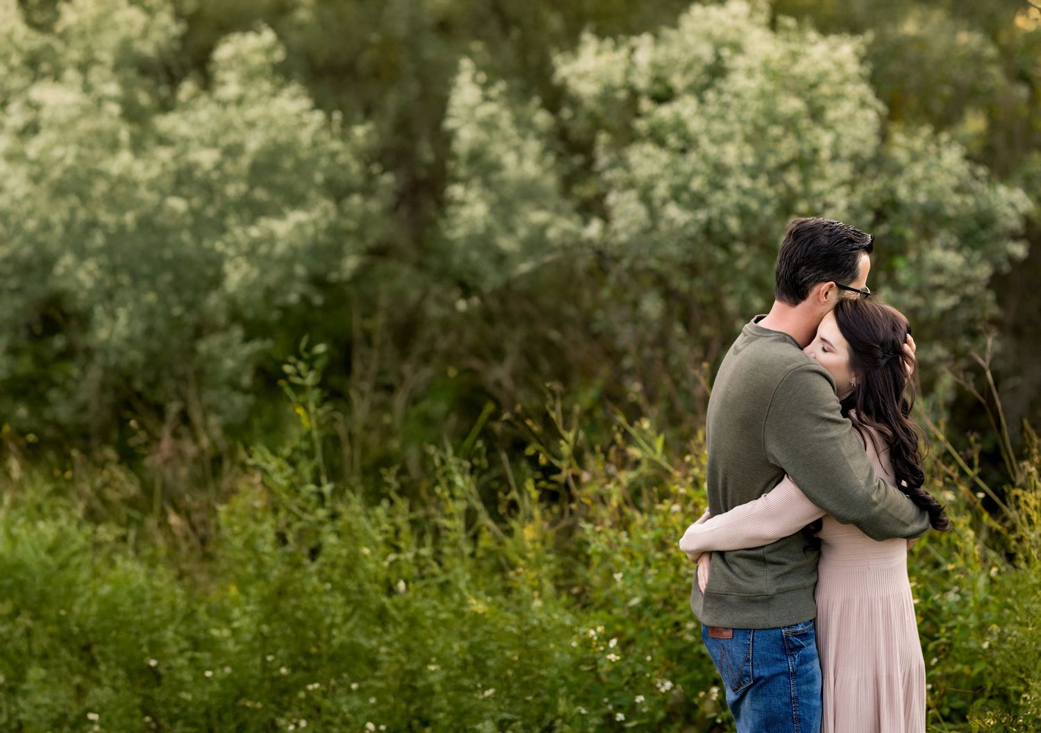 A couple embraces in a romantic moment while standing in a lush green meadow on a sunny day.