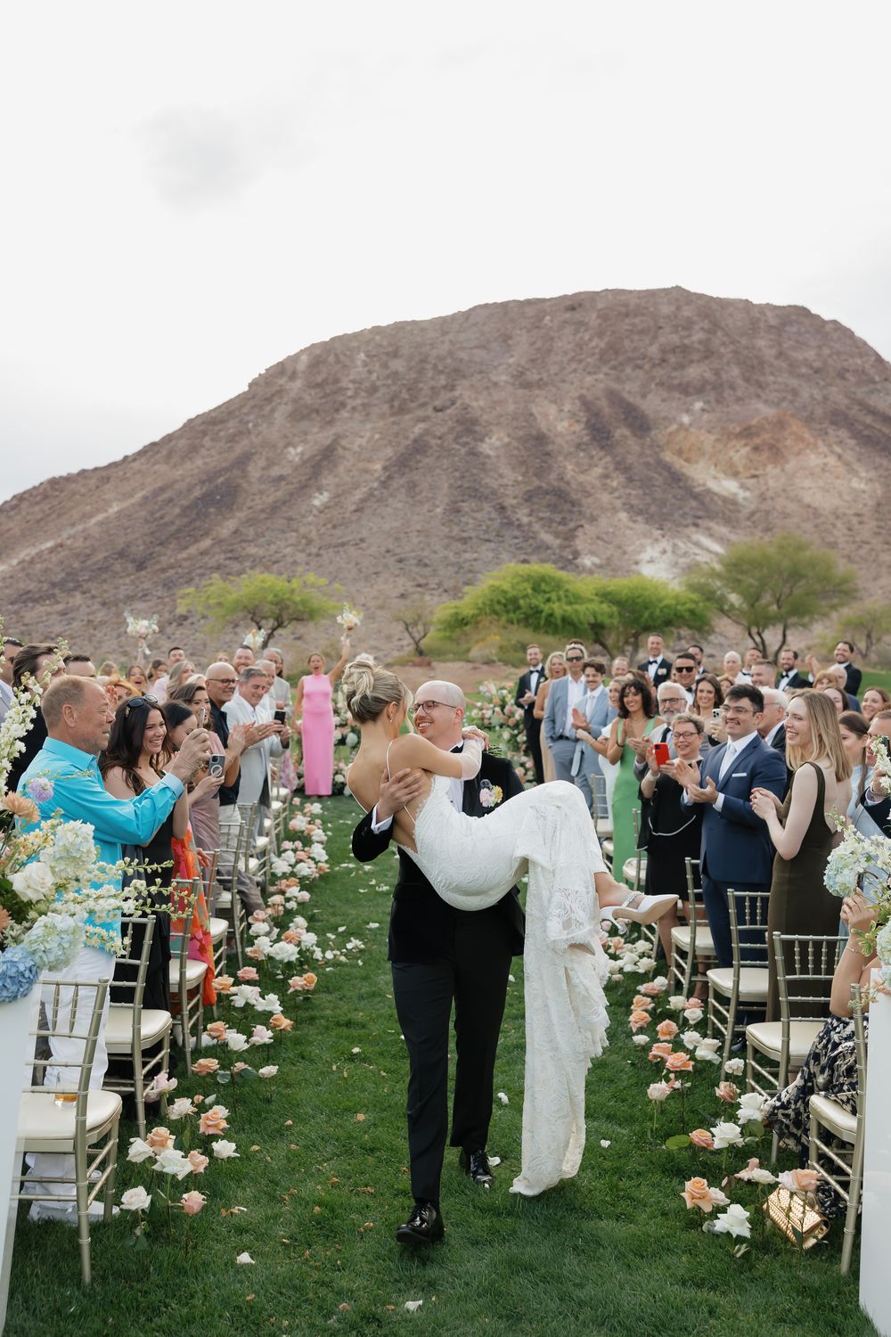 Wedding guests seated in white chairs with floral aisle decorations and mountain backdrop.