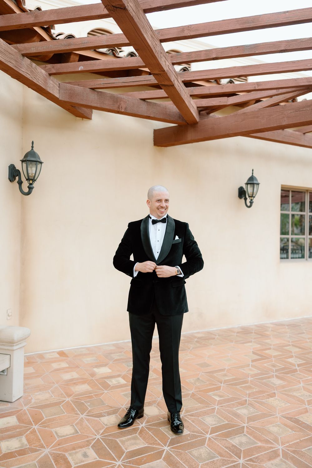 Groom in classic black tuxedo adjusting his outfit under wooden beams.