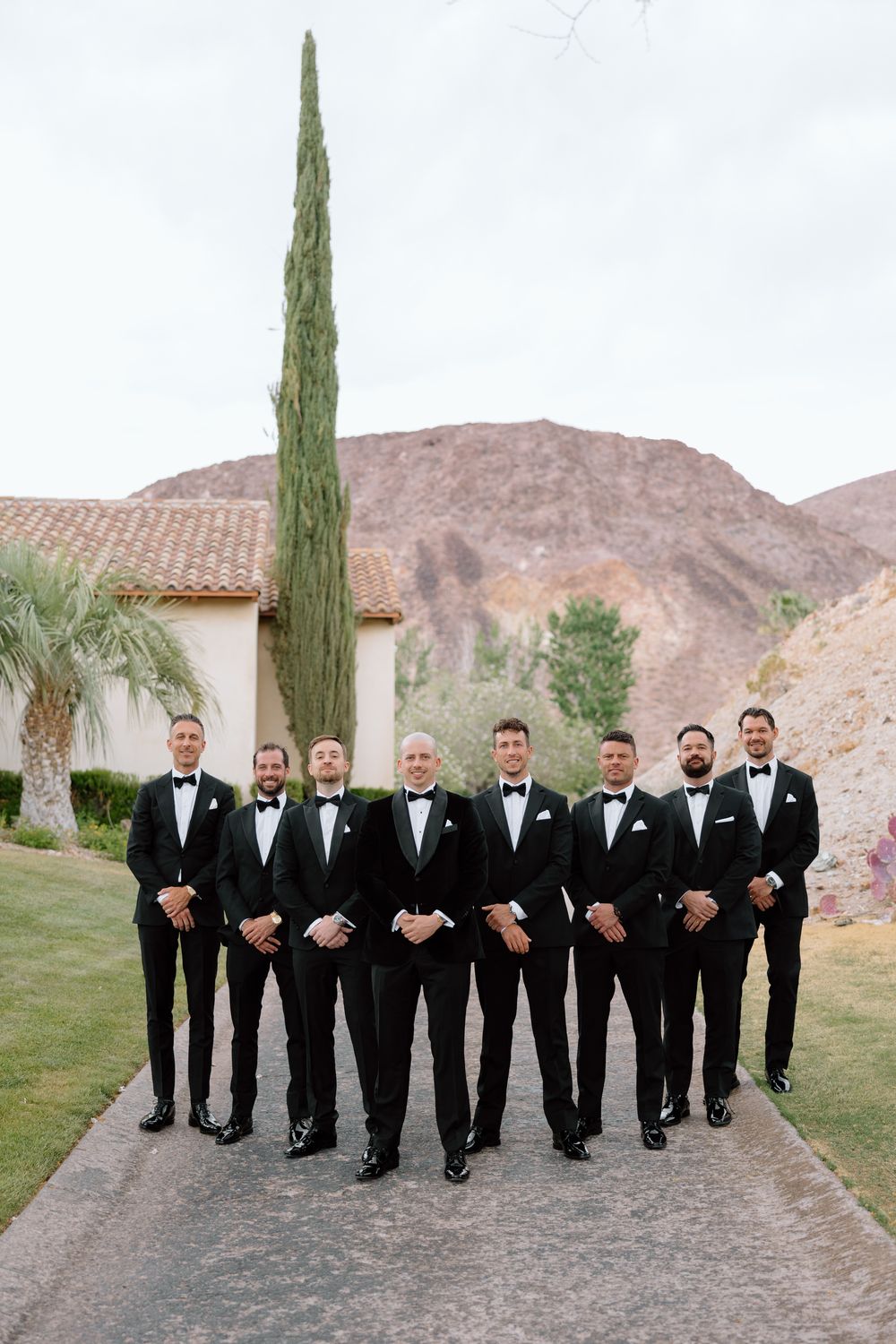 Groomsmen in black tuxedos pose together on path with desert mountains in background.