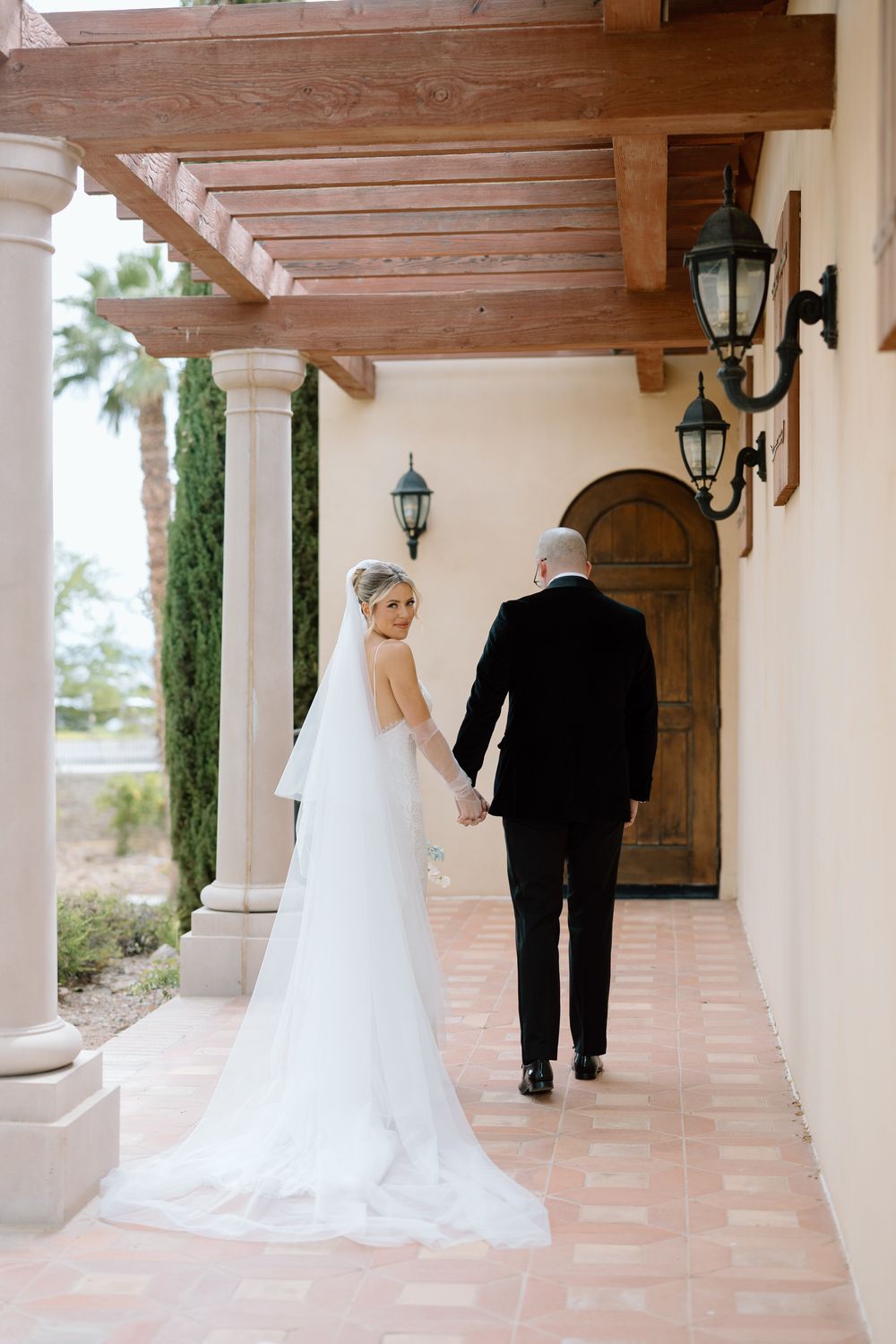Bride and groom walk together along covered terracotta tile walkway with wooden beams.