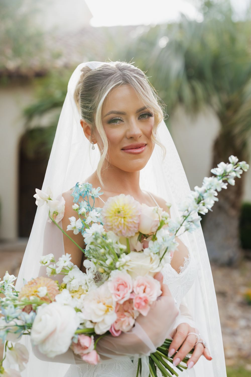 Bride holds pastel bouquet with pink roses and white flowers against garden backdrop.