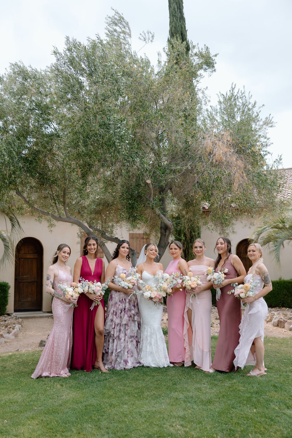 Bridesmaids in pink and floral dresses pose together on grass in front of Mediterranean style building.