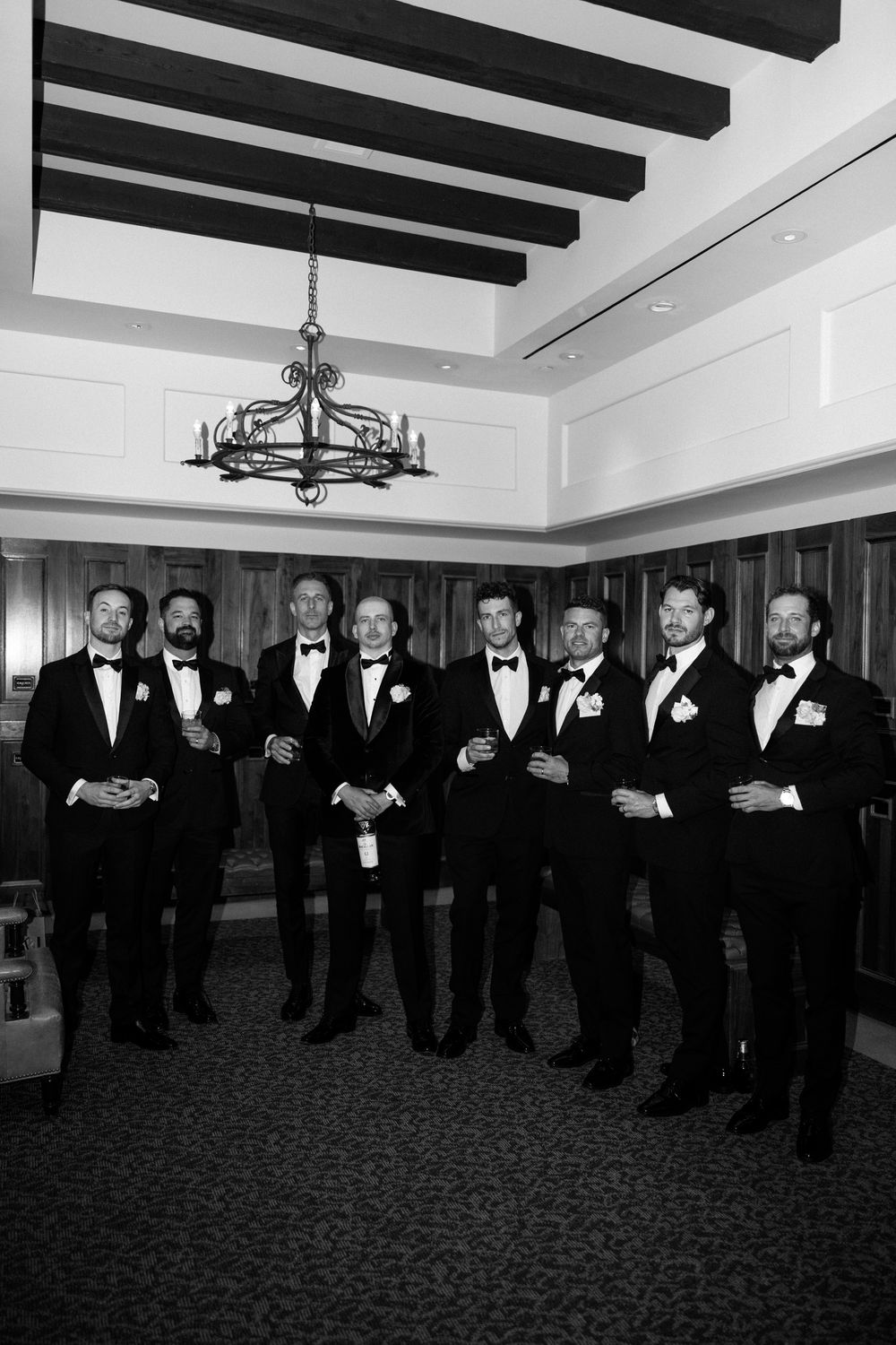 Groomsmen in black tuxedos line up in formation in an elegant room with exposed ceiling beams.