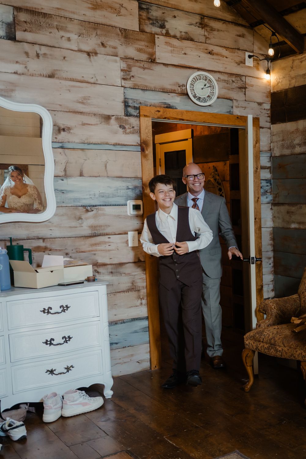 Two people stand beside white vintage furniture in a rustic room with wooden plank walls.