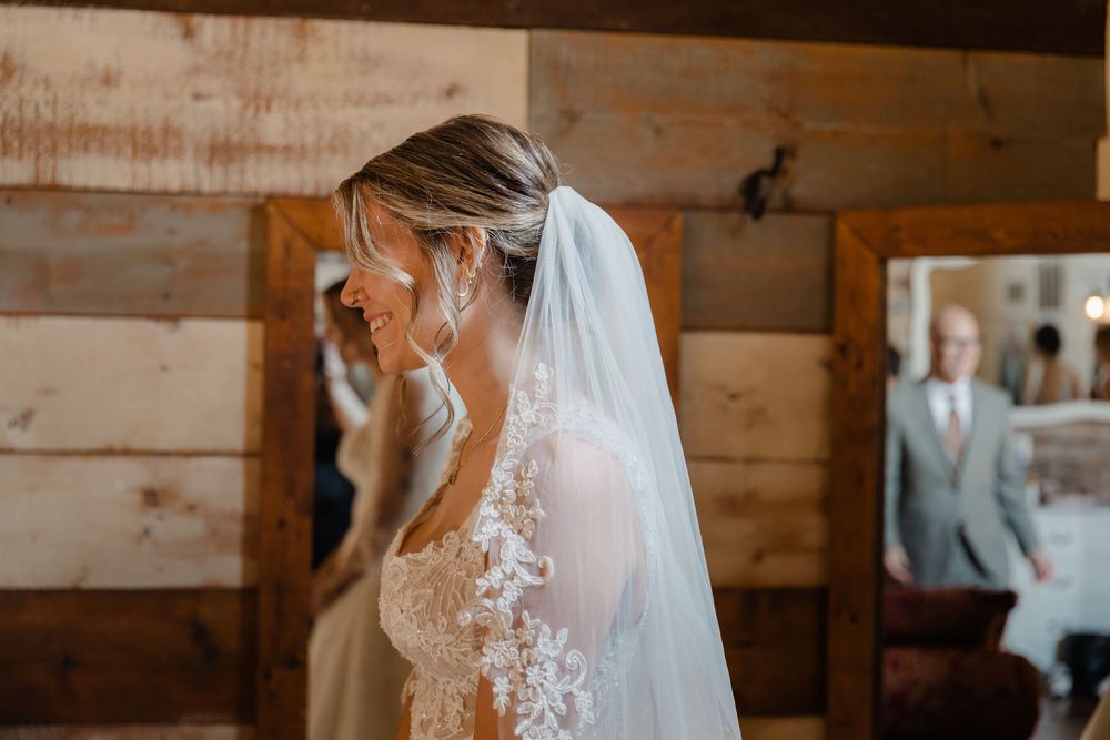 A bride in a delicate lace wedding dress and flowing veil stands in a rustic wooden barn setting.