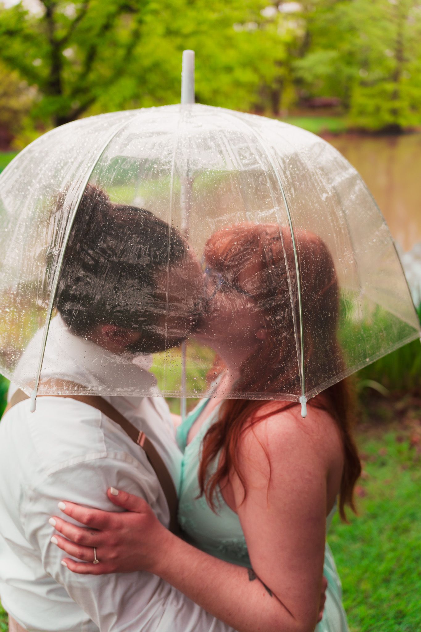 Couple shares intimate moment under clear umbrella in lush green garden setting.