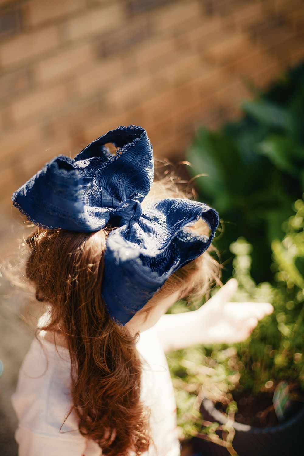 Young girl with large navy blue velvet bow in curly red hair standing near brick wall.