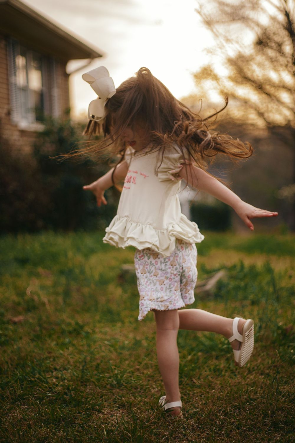 A young girl in a white ruffled top and floral shorts twirls joyfully in a backyard at golden hour.