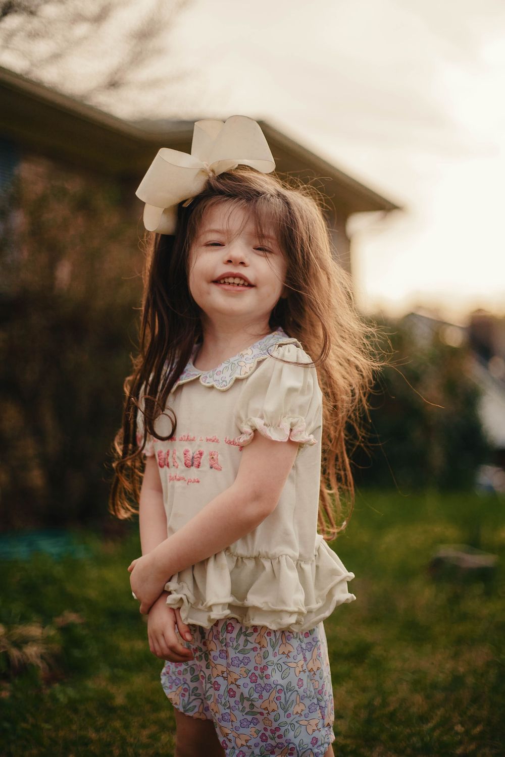A smiling little girl with a large cream bow in her hair wearing a white ruffled top and floral shorts outdoors.