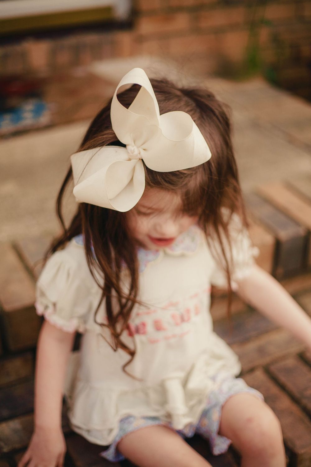 A little girl with long brown hair wearing a large cream bow headband sits on wooden steps outdoors.