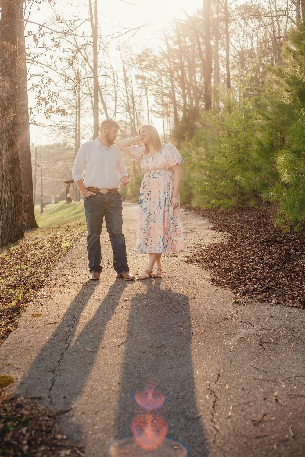 Couple holding hands walking on sunlit path surrounded by tall pine trees at golden hour.