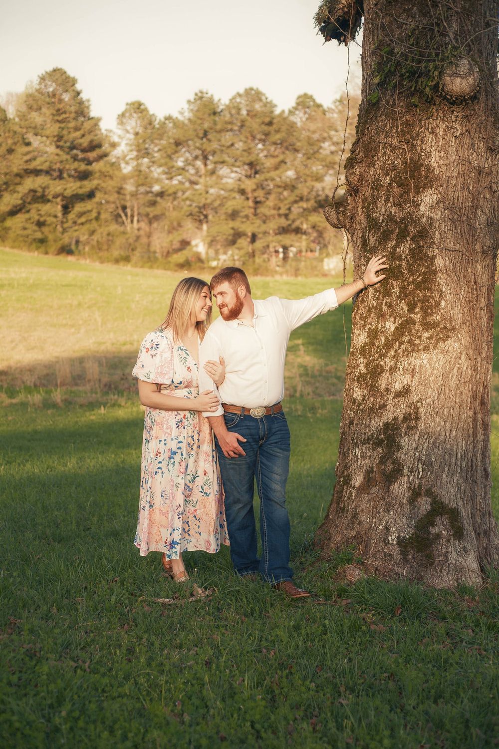 Couple embracing near large tree trunk in lush green field during warm sunset engagement session.
