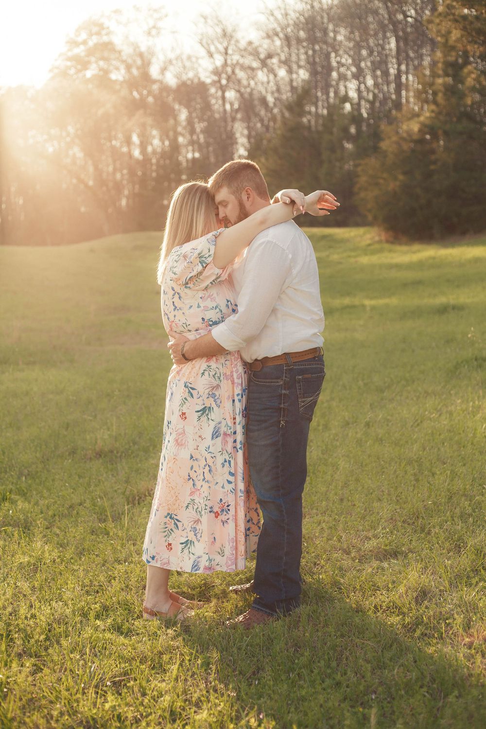 Couple sharing tender embrace in open green meadow bathed in golden sunset light.