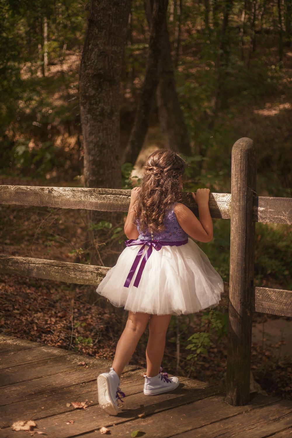 Flower girl in white tutu and purple bow dress standing on wooden bridge in forest setting.