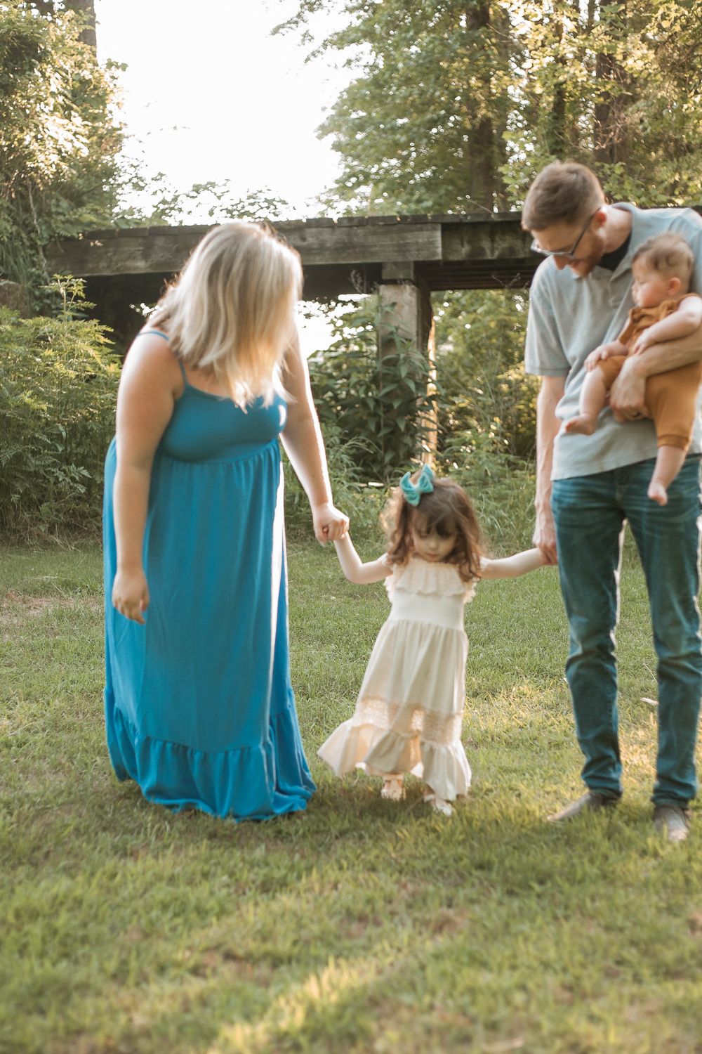 Family of four holding hands on green grass near rustic wooden bridge in summer park.