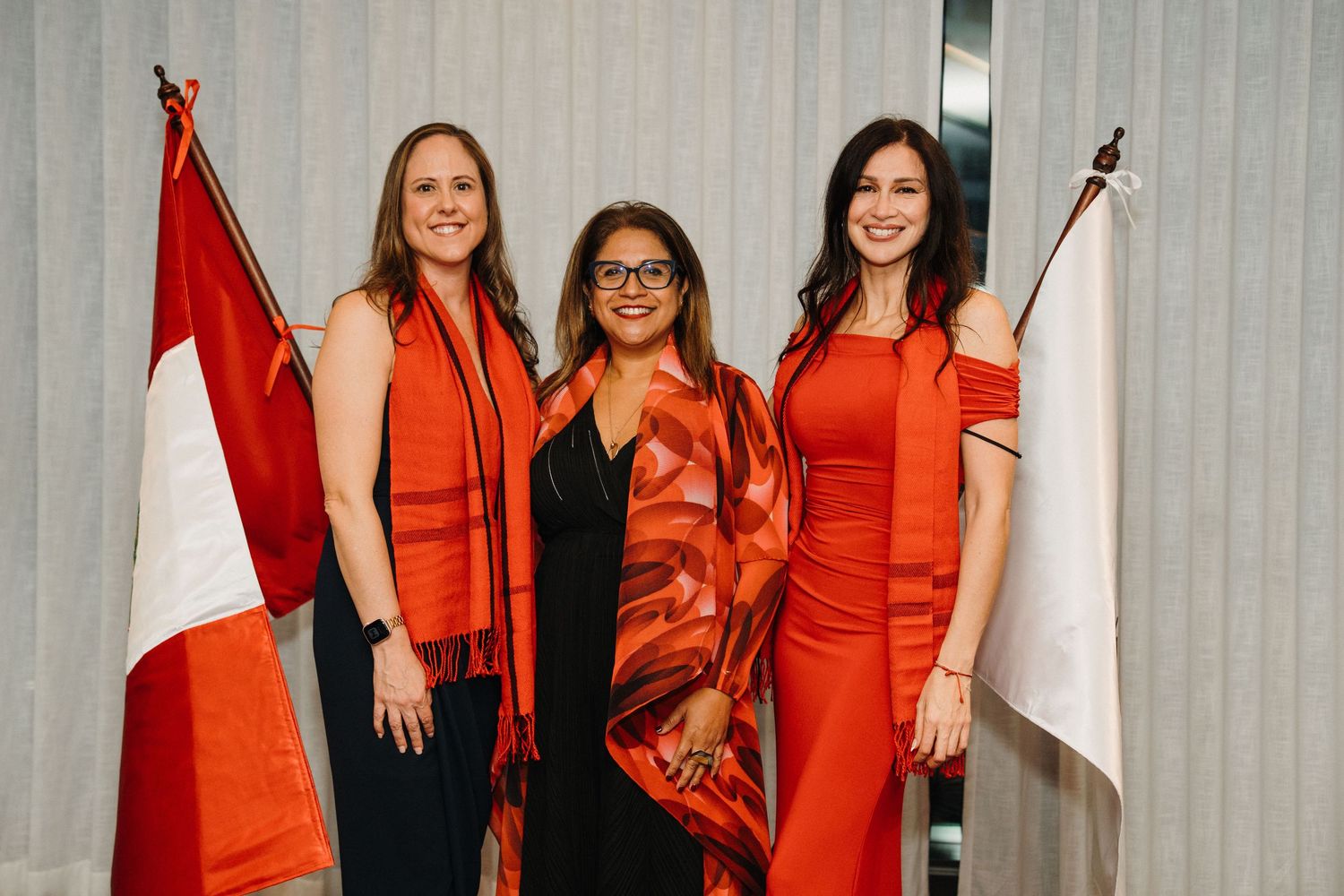 Three people in orange outfits stand together holding Canadian flags against a light background.
