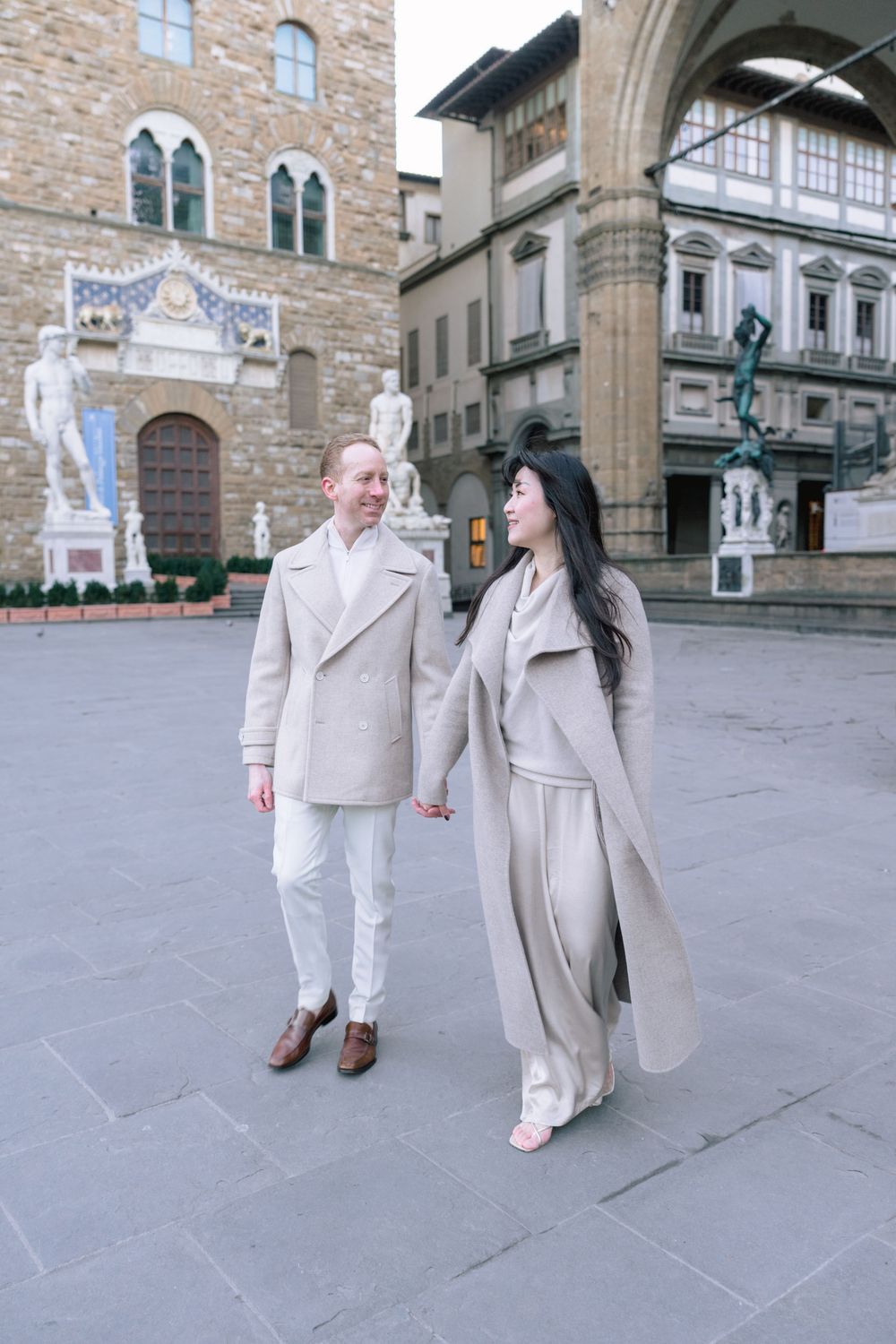 A couple in coordinating light-colored outfits strolls together on a stone plaza in front of historic Italian architecture.