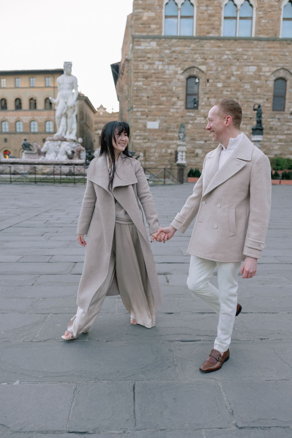 A couple in beige and white outfits dancing together on a stone plaza outside a historic building.